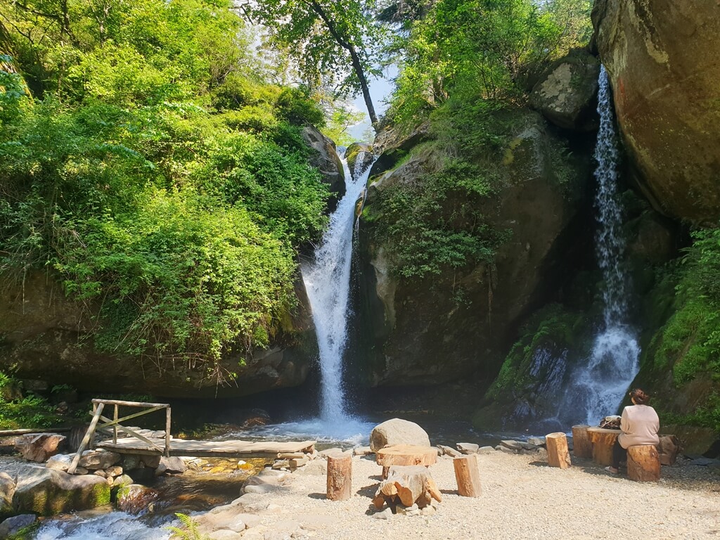 The twin water streams of the Sajla waterfall with an open seating arrangement on either side connected by a wooden bridge